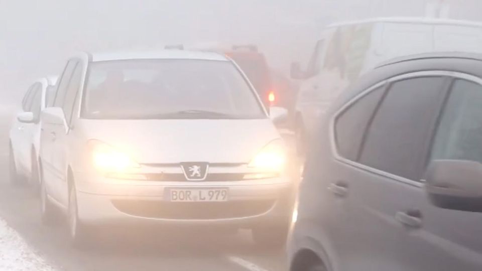 Im Nebel stauen sich auf einer Straße Autos mit Licht an. Neben der Straße liegt Schnee