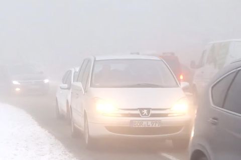 Im Nebel stauen sich auf einer Straße Autos mit Licht an. Neben der Straße liegt Schnee