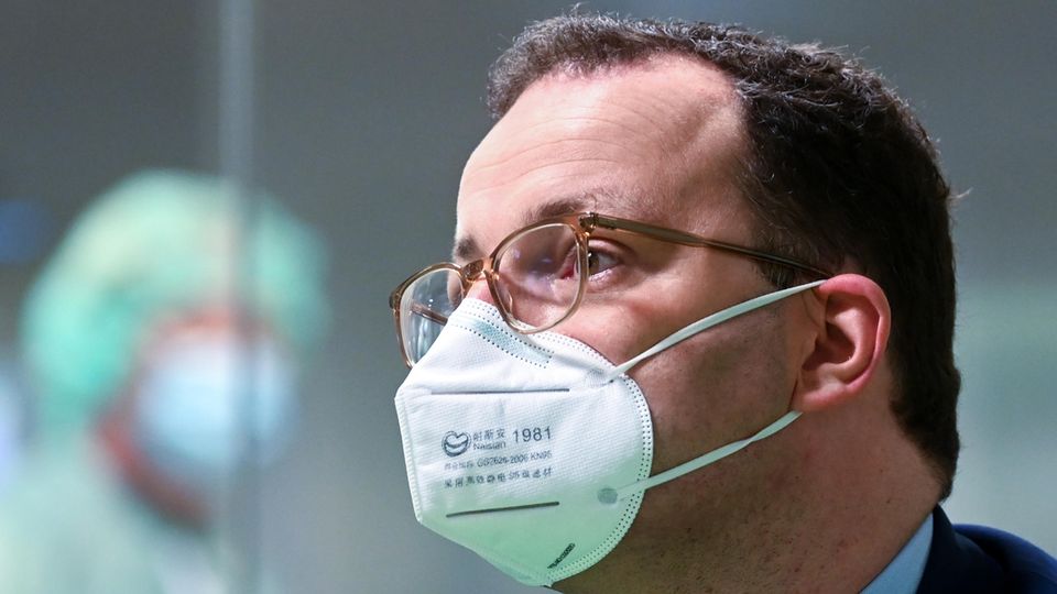 Bundesgesundheitsminister Spahn mit FFP2-Maske Ein Mann mit braunen Locken trägt eine FFP2-Maske sowie einen blauen Anzug. Im Hintergrund ist schemenhaft ein Labor zu sehen