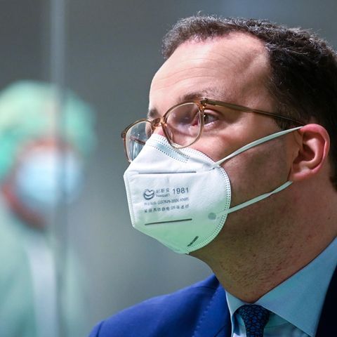 Ein Mann mit braunen Locken trägt eine FFP2-Maske sowie einen blauen Anzug. Im Hintergrund ist schemenhaft ein Labor zu sehen