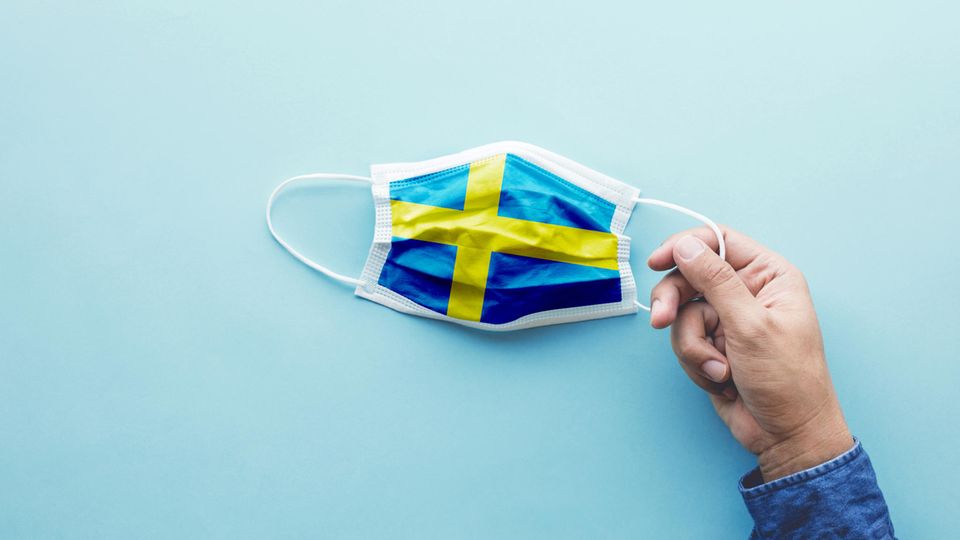 Eine Hand hält einen Mund-Nase-Schutz mit der schwedischen Flagge als Motiv