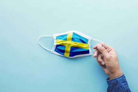 Eine Hand hält einen Mund-Nase-Schutz mit der schwedischen Flagge als Motiv
