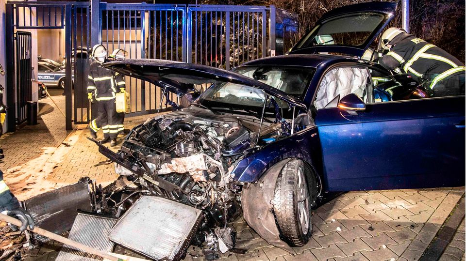 Feuerwehrleute stehen neben einem PKW, der gegen die Torausfahrt einer Polizeidienststelle geprallt ist
