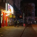 Vor einem Supermarkt mit Leuchtreklame im Stil eines Stripclubs gehen zwei junge Männer mit dricken Jacken und Rucksäcken vorbei