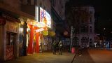Vor einem Supermarkt mit Leuchtreklame im Stil eines Stripclubs gehen zwei junge Männer mit dricken Jacken und Rucksäcken vorbei