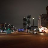Eine rot asphaltierter Platz liegt in der Dunkelheit leer da. Links eine vierspurige Straße, rechts spärlich beleuchtete Häuser