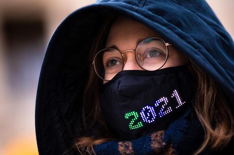 Eine Frau mit Brille trägt eine dunkelblaue Kapuze sowie einen schwarzen Mund-Nasen-Schutz mit bunter "2021"-Stickerei
