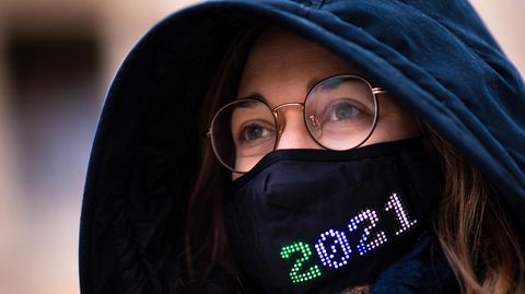 Eine Frau mit Brille trägt eine dunkelblaue Kapuze sowie einen schwarzen Mund-Nasen-Schutz mit bunter "2021"-Stickerei