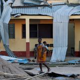 Eine Frau trägt ein Baby auf ihrem Rücken und geht an einer beschädigten Schule vorbei.